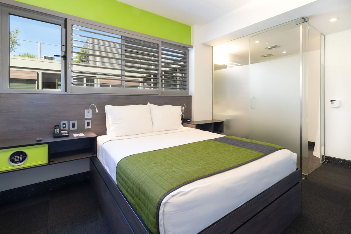A modern hotel room with a bed, a nightstand, a green accent wall, a window with blinds, a glass shower enclosure, and a view of the outside.
