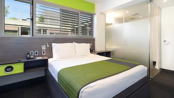 A modern hotel room with a bed, a nightstand, a green accent wall, a window with blinds, a glass shower enclosure, and a view of the outside.