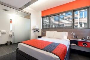 A hotel room with a bed, bedside table, and a window with blinds.