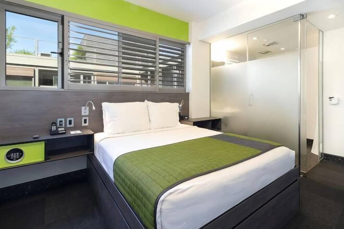 A modern hotel room with a bed, bedside table, and a window with blinds.