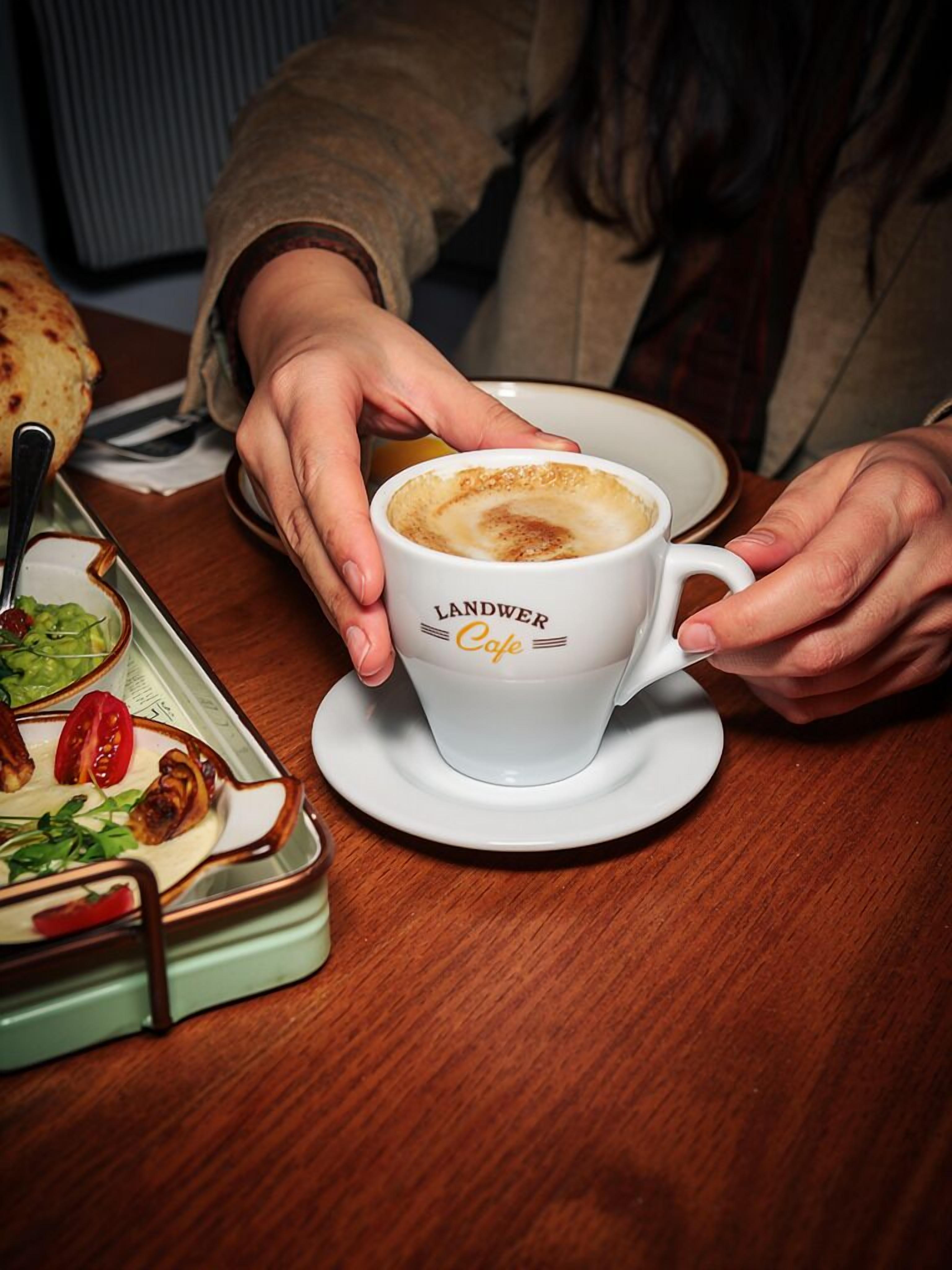 Eine Person hält eine Kaffeetasse von Landwer Cafe, während auf dem Tisch ein Teller mit Speisen und ein Brötchen stehen.
