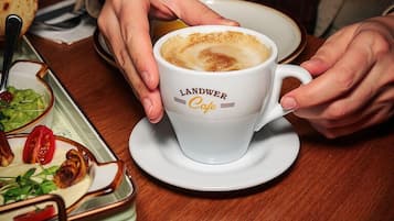 Eine Person hält eine Kaffeetasse von Landwer Cafe, während auf dem Tisch ein Teller mit Speisen und ein Brötchen stehen.
