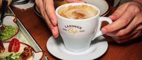 Una persona sosteniendo una taza de café de Landwer Cafe, con un plato de comida y un panecillo sobre la mesa.