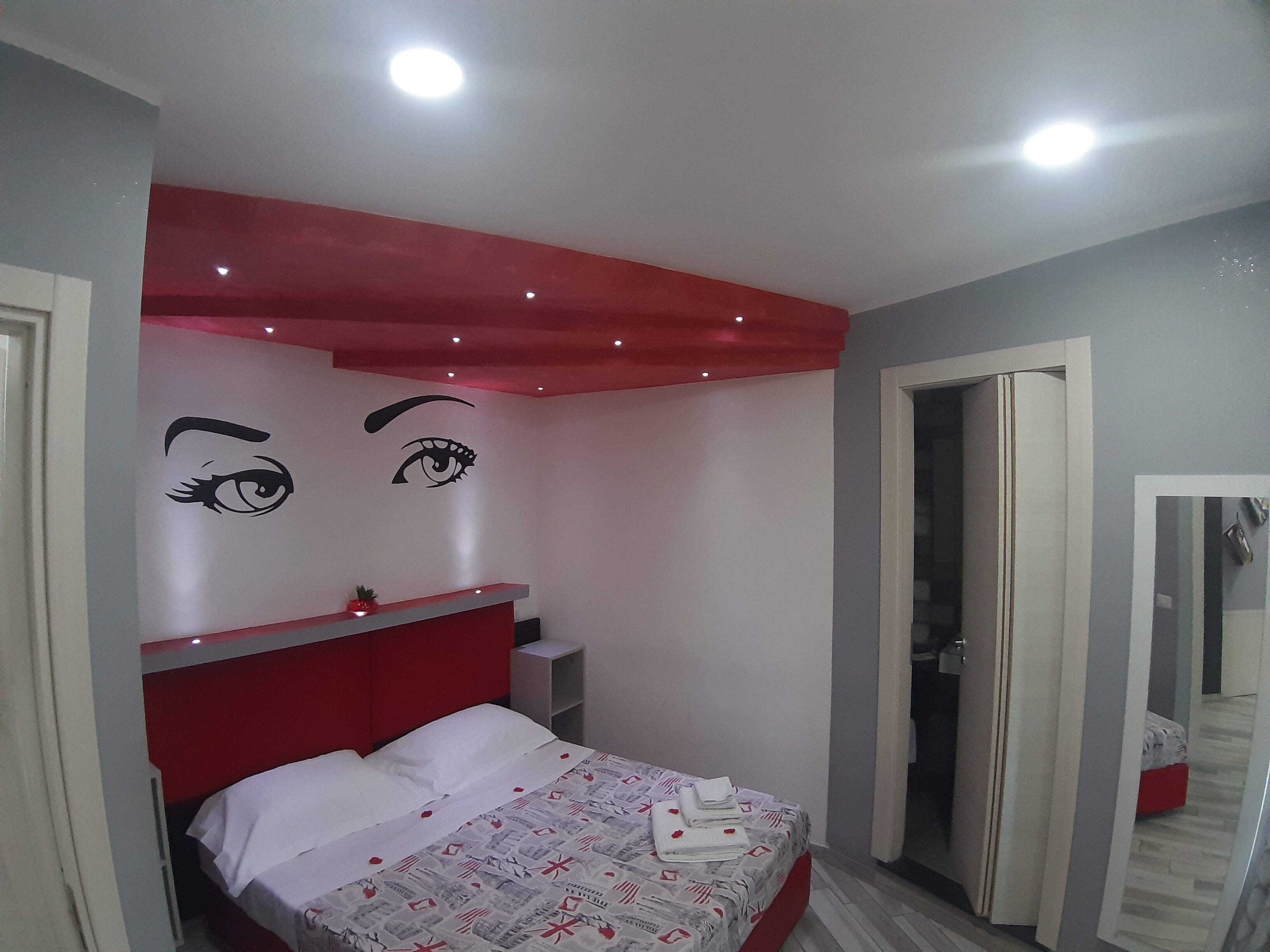 A modern bedroom with a red headboard, a bed with white linens, and a wall with two large eye designs.