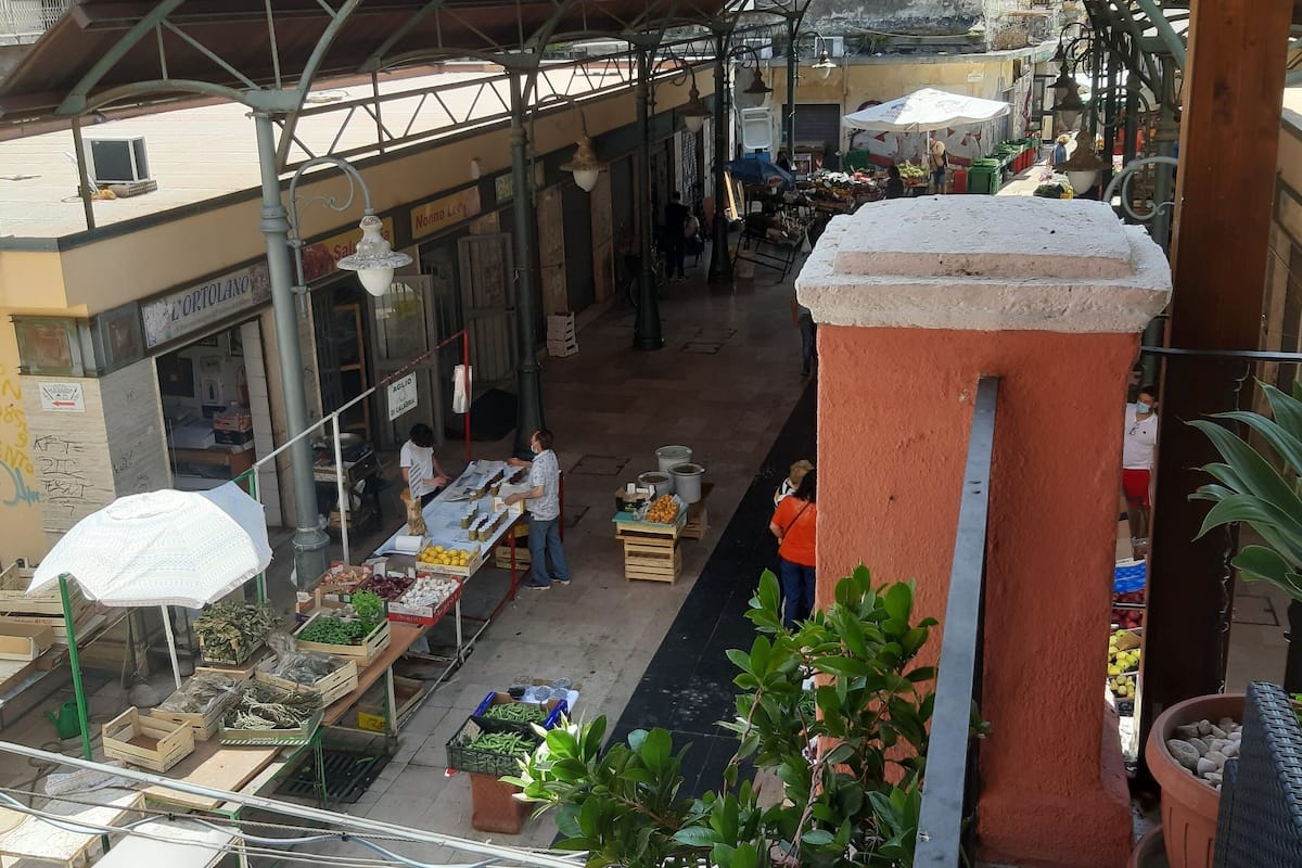 An outdoor market with vendors and customers, covered by a metal roof structure.