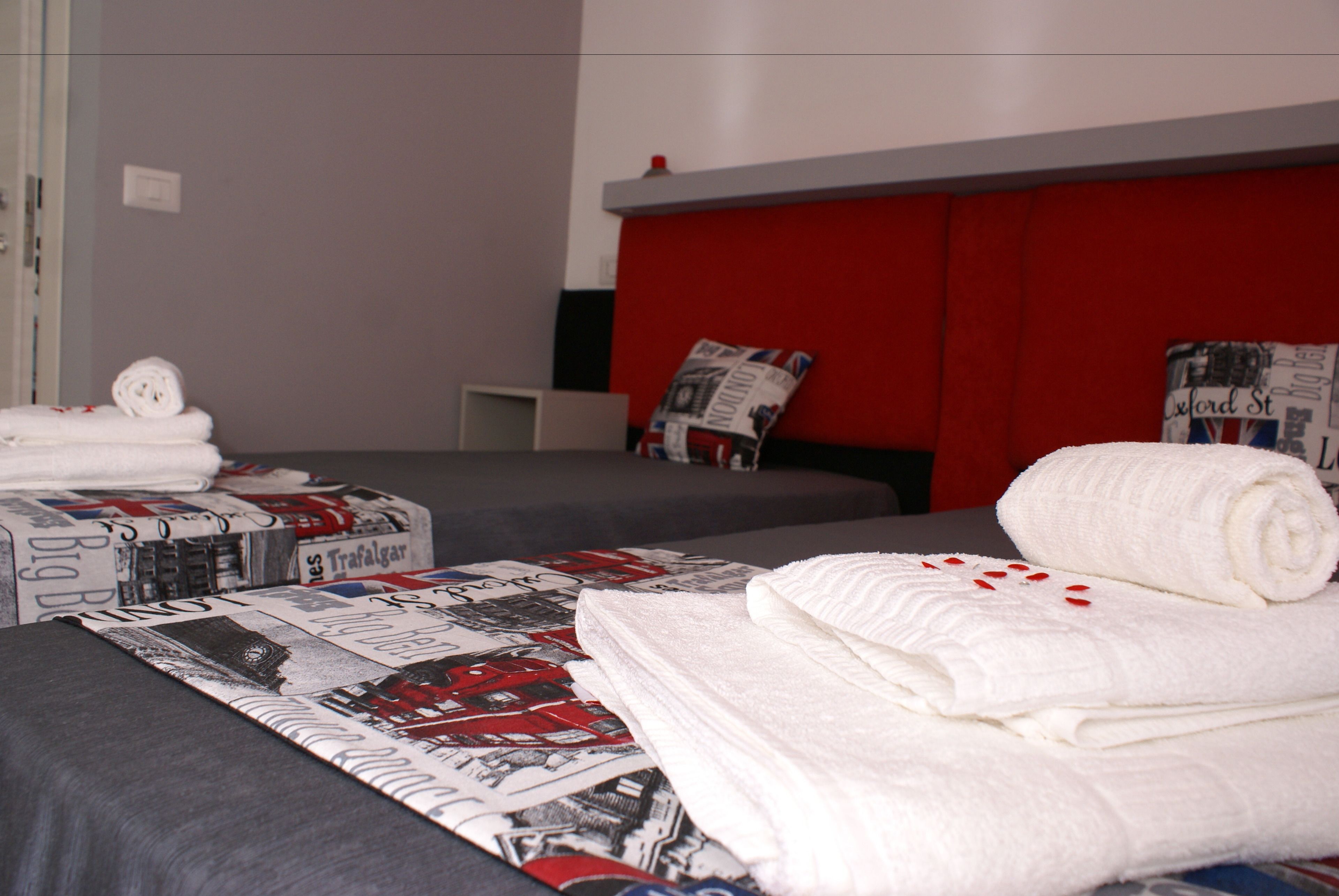 Quarto de hotel com duas camas, uma com cabeceira vermelha e outra com cabeceira cinza. Toalhas brancas dobradas sobre as camas.