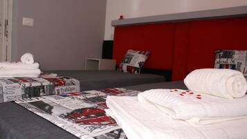 Quarto de hotel com duas camas, uma com cabeceira vermelha e outra com cabeceira cinza. Toalhas brancas dobradas sobre as camas.