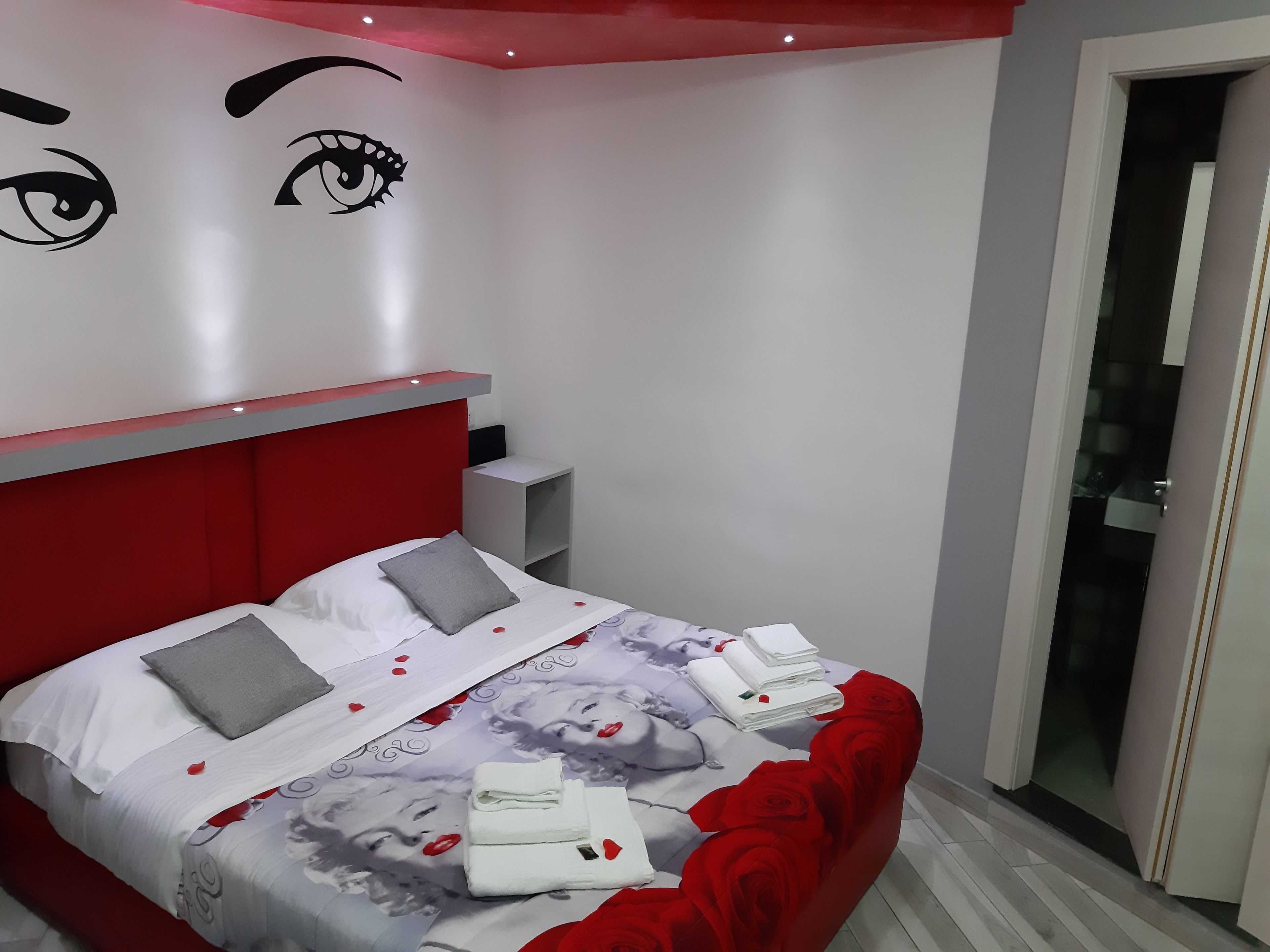 A modern bedroom with a red headboard, a bed with white bedding and red rose decorations, a nightstand, and a wall with a large eye design.