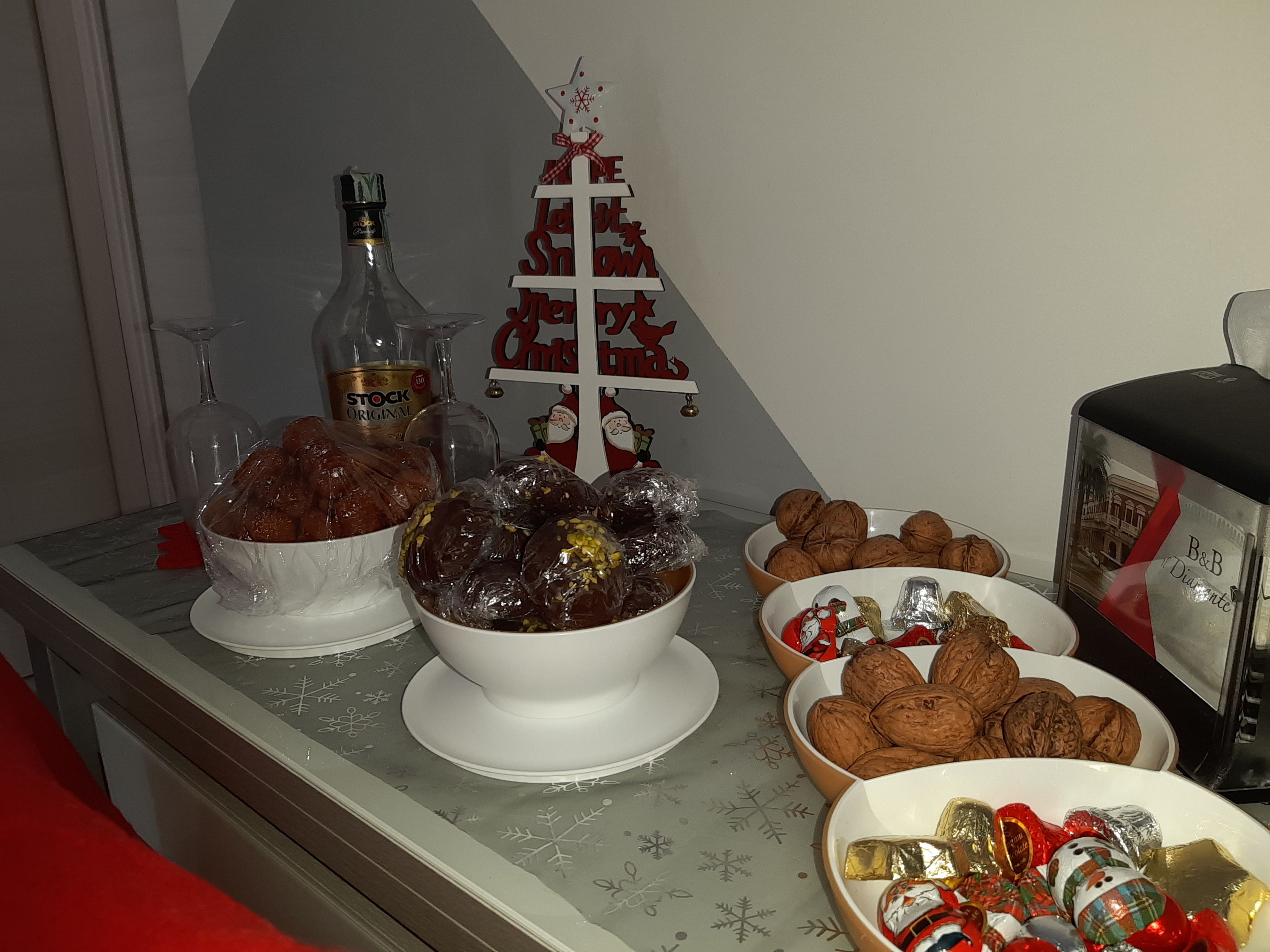 A buffet table with various food items, including wrapped dates, nuts, and chocolates, set against a backdrop with a small Christmas tree and a bottle of liquor.