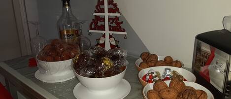 Uma mesa de buffet com diversos alimentos, incluindo tâmaras envoltas, nozes e chocolates, com um pequeno pinheiro de Natal e uma garrafa de licor ao fundo.
