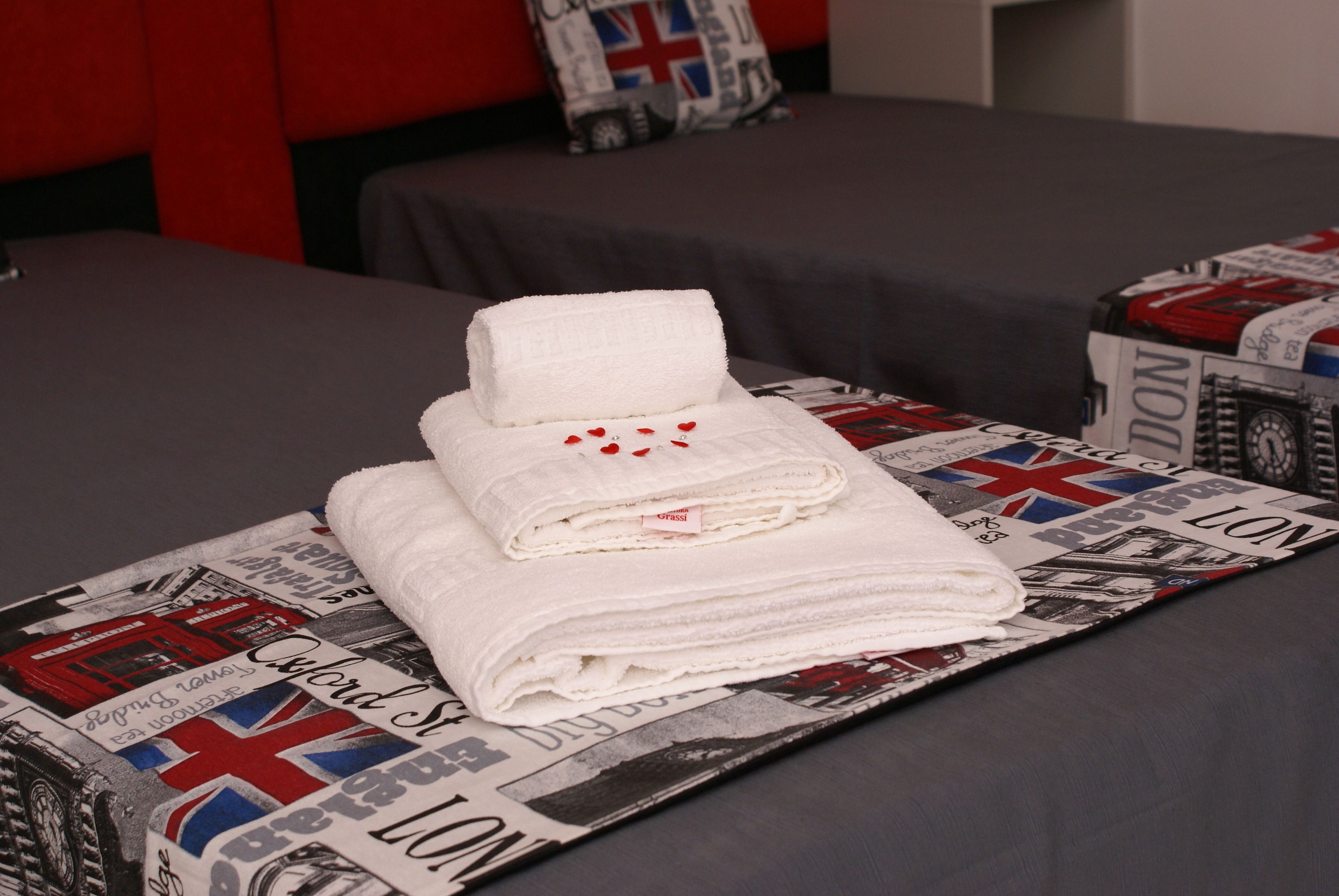 A neatly made bed with a folded white towel and a rolled towel on top, placed on a bedspread with a newspaper print design.