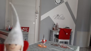 Una habitación con pared blanca, un reloj, un pequeño árbol de Navidad y una mesa con mantel rojo y un objeto con forma de corazón rojo.