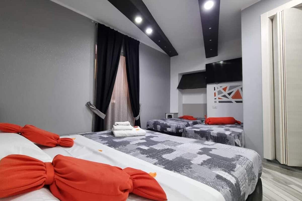 A hotel room with two beds, one with a white and orange bedspread and the other with a grey and white patterned bedspread. There is a small table with a red pillow and a white towel on it. The room has a grey wall and a black ceiling with recessed lighting.