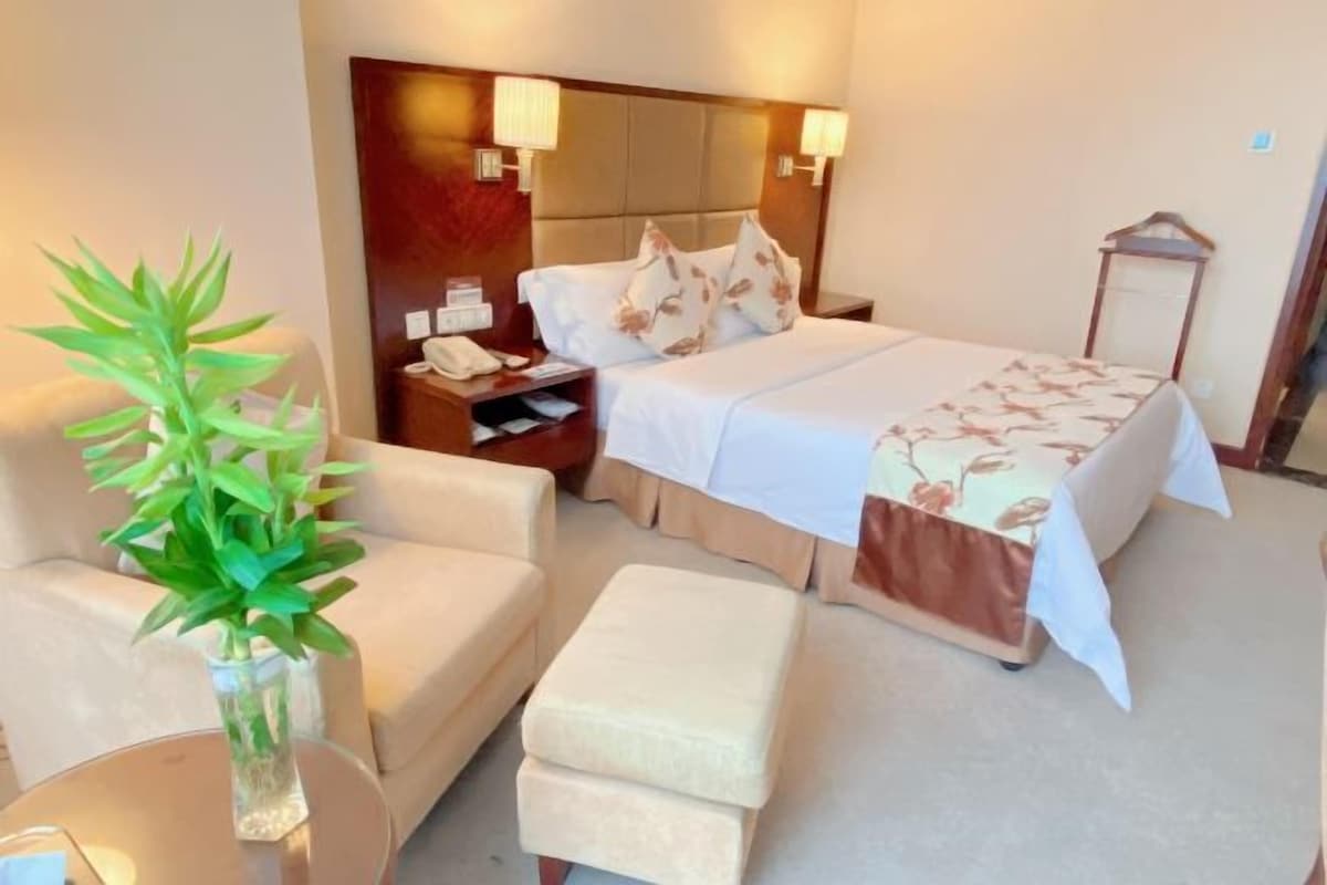 Una habitación de hotel con cama, mesita de noche, lámpara, sillón y planta en maceta.