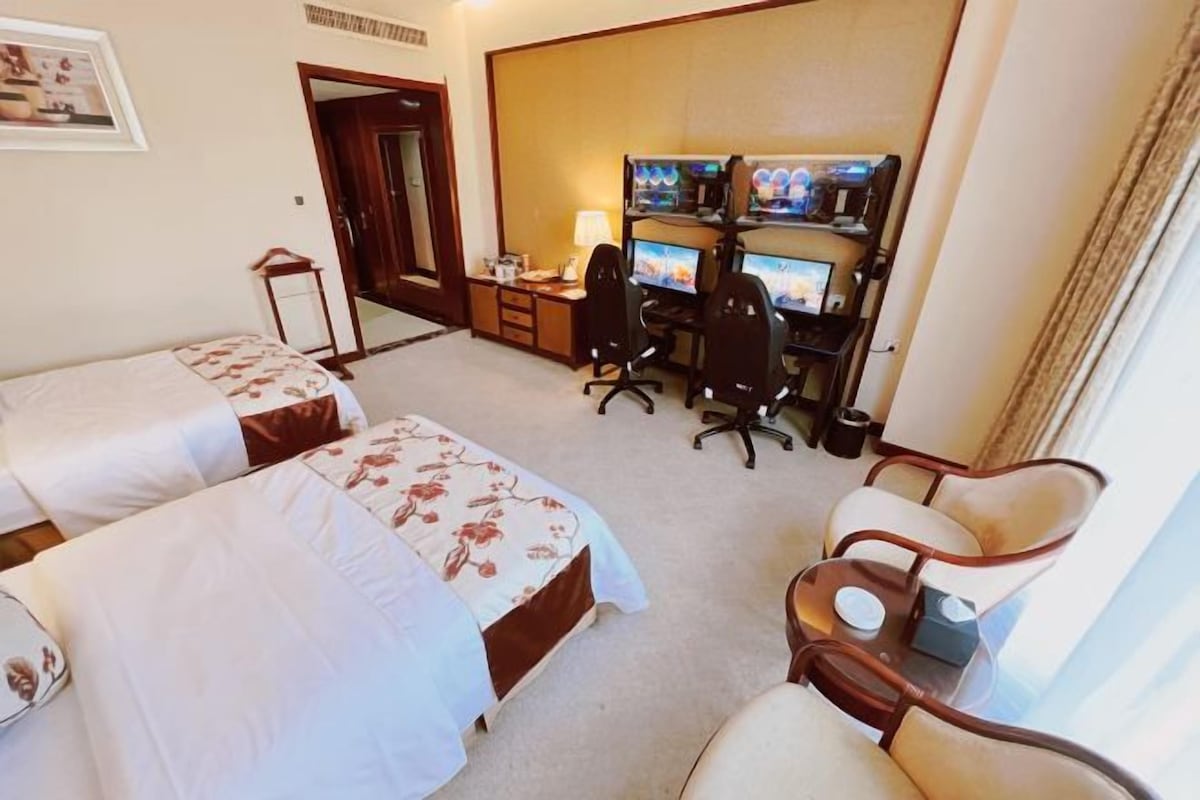 Habitación de hotel con dos camas, un escritorio con varios monitores, una silla y una mesita.