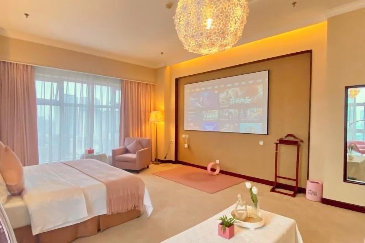 Habitación de hotel con cama, escritorio, silla, pantalla de proyector y un amplio ventanal con cortinas.