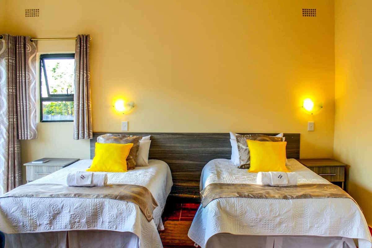 A hotel room with two beds, each with a white bedspread and a pillow. There are two bedside tables with lamps, a window with curtains, and a wall-mounted light fixture.