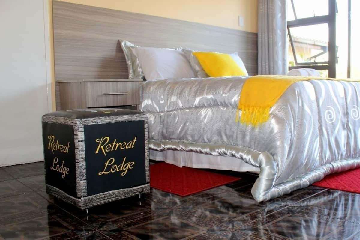 A neatly made bed with a grey and white patterned bedspread, a red rug, and a black side table labeled "Retreat Lodge."