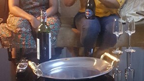 Two women seated on a couch with a table in front of them. The table has a bottle, a glass, and a tray.