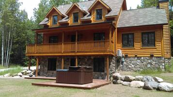 A two-story log cabin with a balcony, stone foundation, and a hot tub in the front yard.