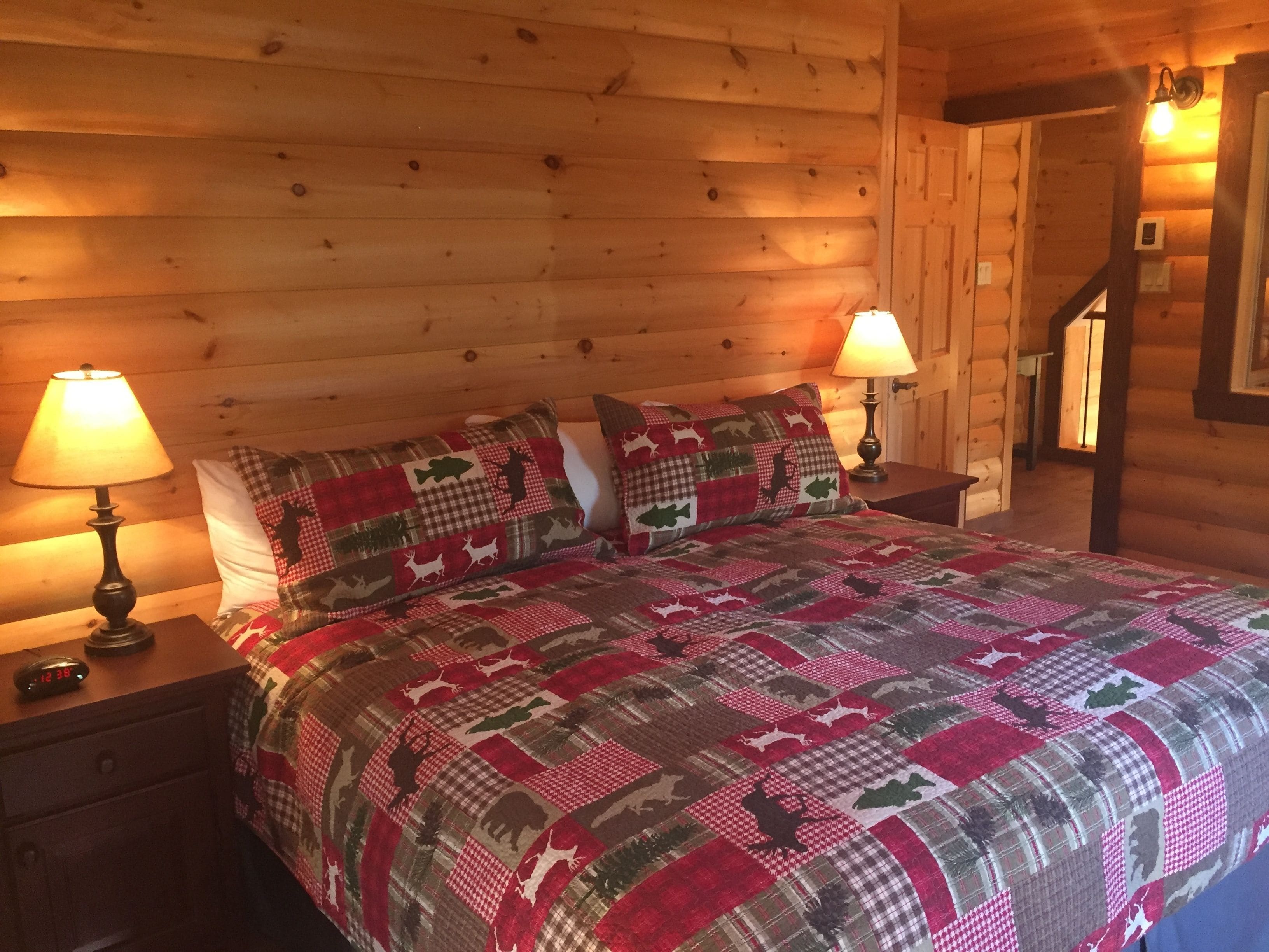 A cozy log cabin bedroom with a wooden headboard, a quilted bedspread, two bedside lamps, and a nightstand with a clock.