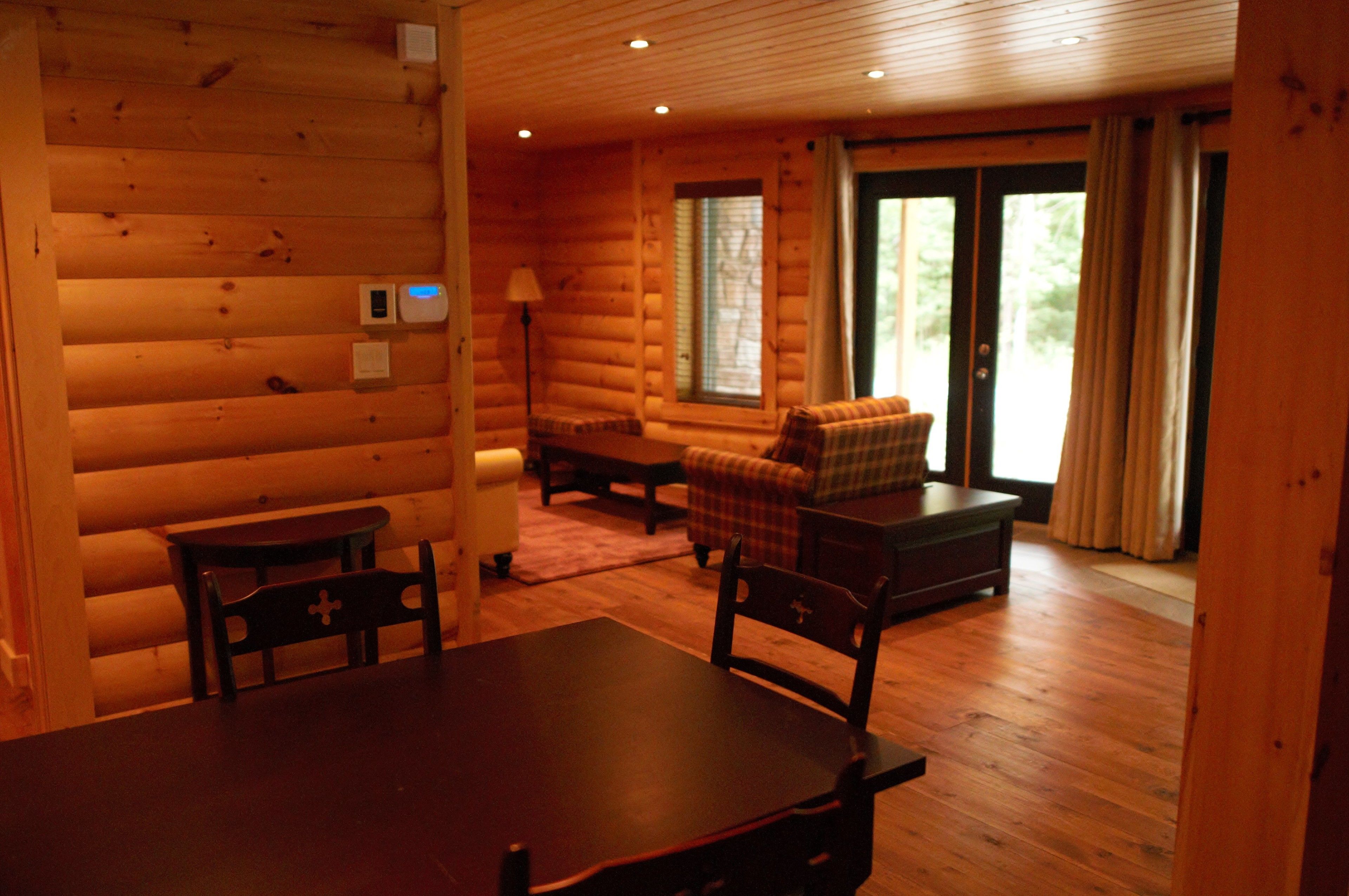 Interior de una cabaña de troncos con una mesa de comedor, sillas, un sofá y una ventana con cortinas.