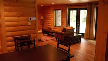 Interior de una cabaña de troncos con una mesa de comedor, sillas, un sofá y una ventana con cortinas.