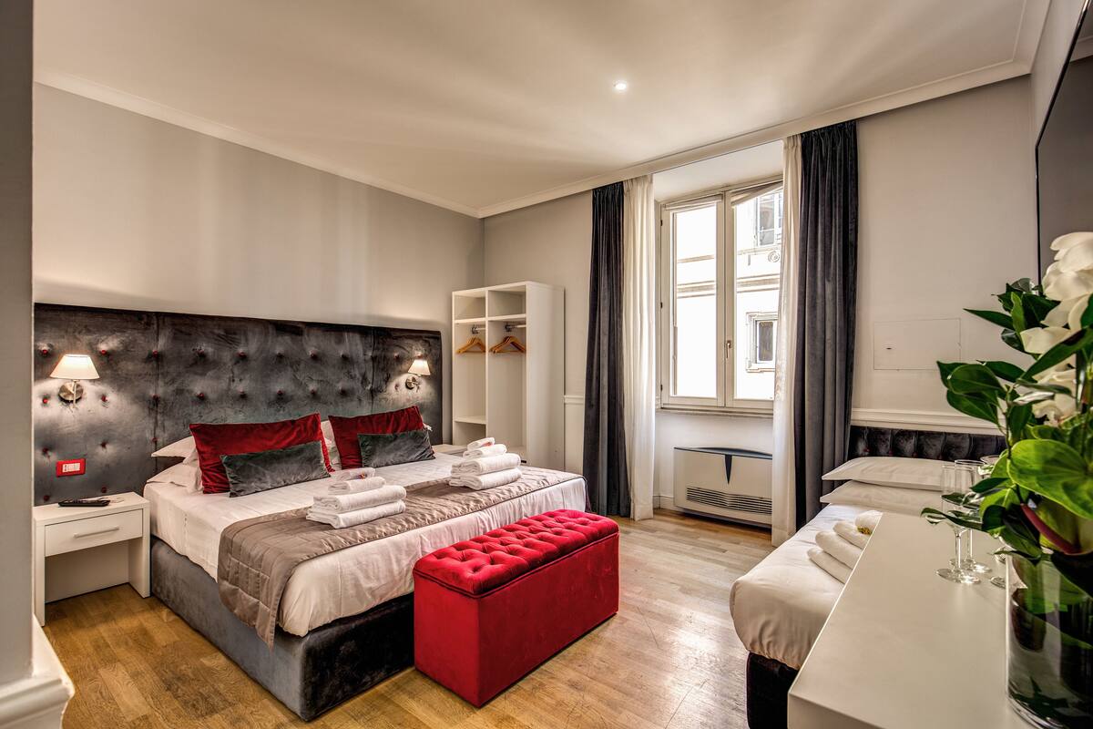 A modern hotel room with a large bed, a red ottoman, a white desk, and a view of the city through the window.