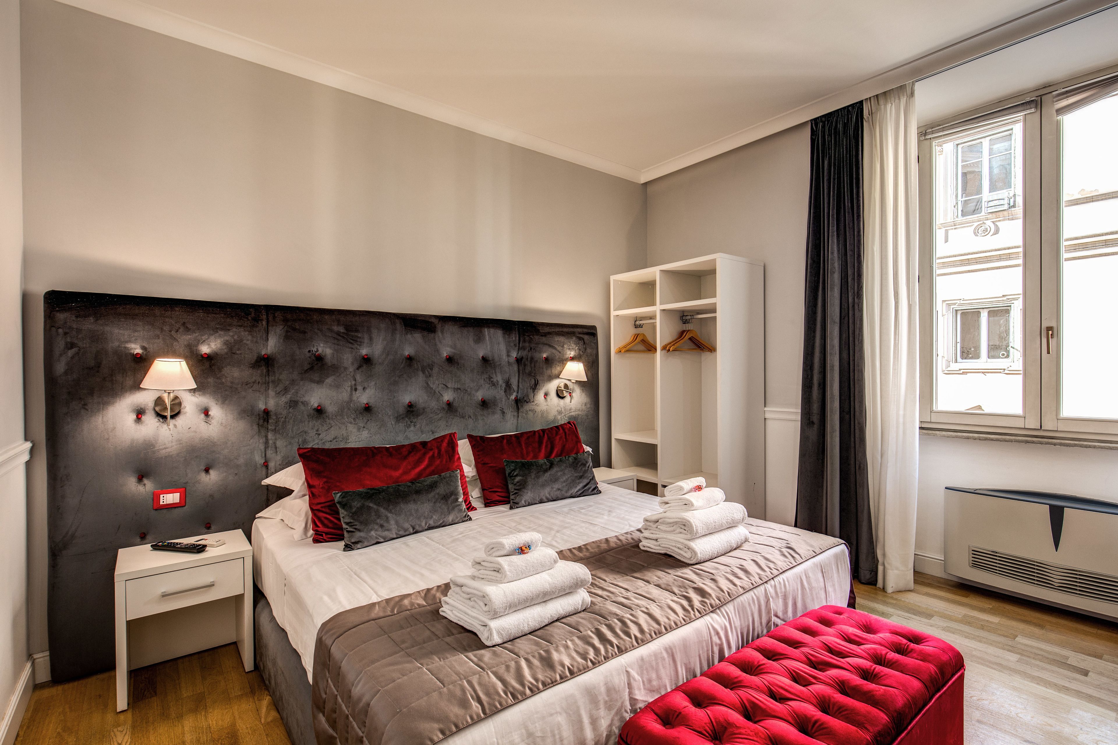 A hotel room with a large bed, a red upholstered bench, a white bedside table, and a wardrobe.