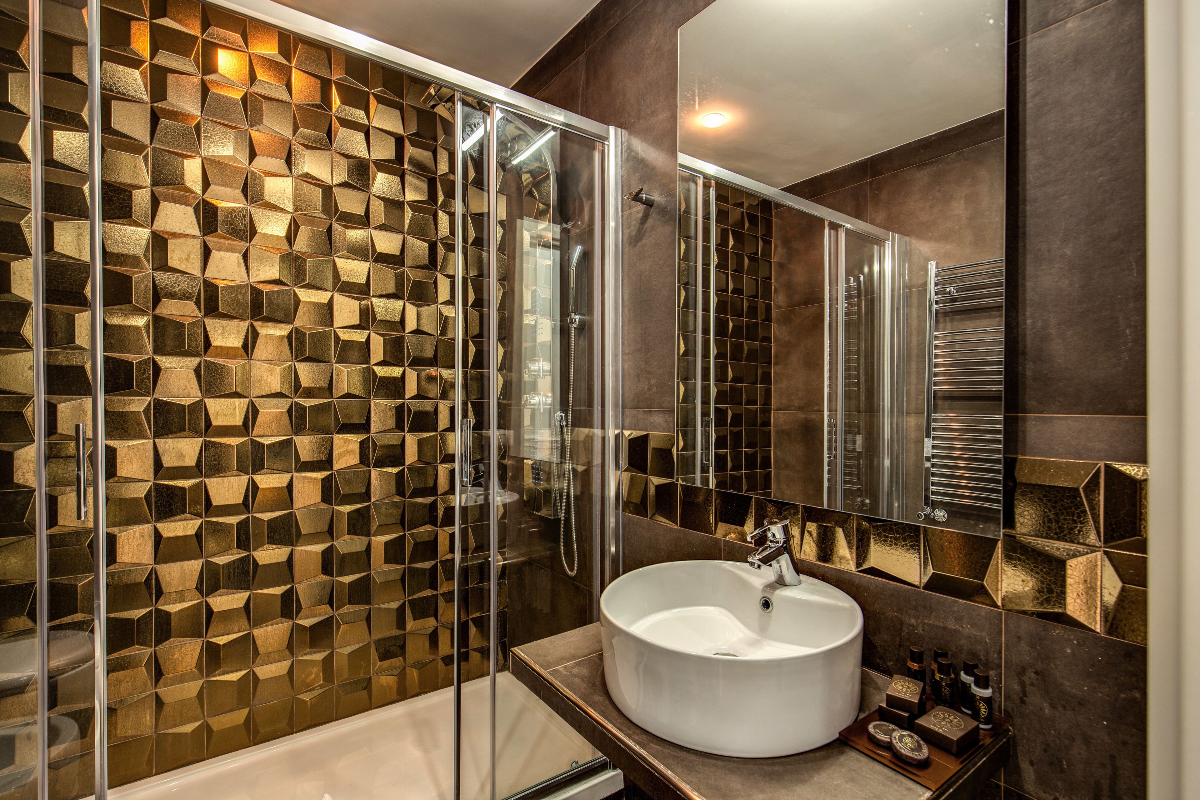 Um banheiro moderno com parede estampada em padrão geométrico, chuveiro com box de vidro e pia redonda com espelho acima.