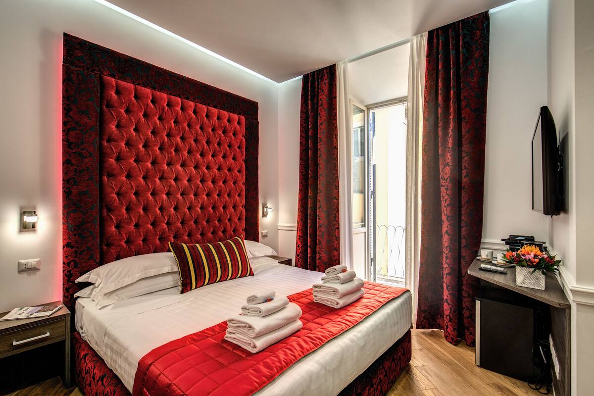 A hotel room with a large bed, red curtains, a flat-screen TV, and a small desk with a vase of flowers.