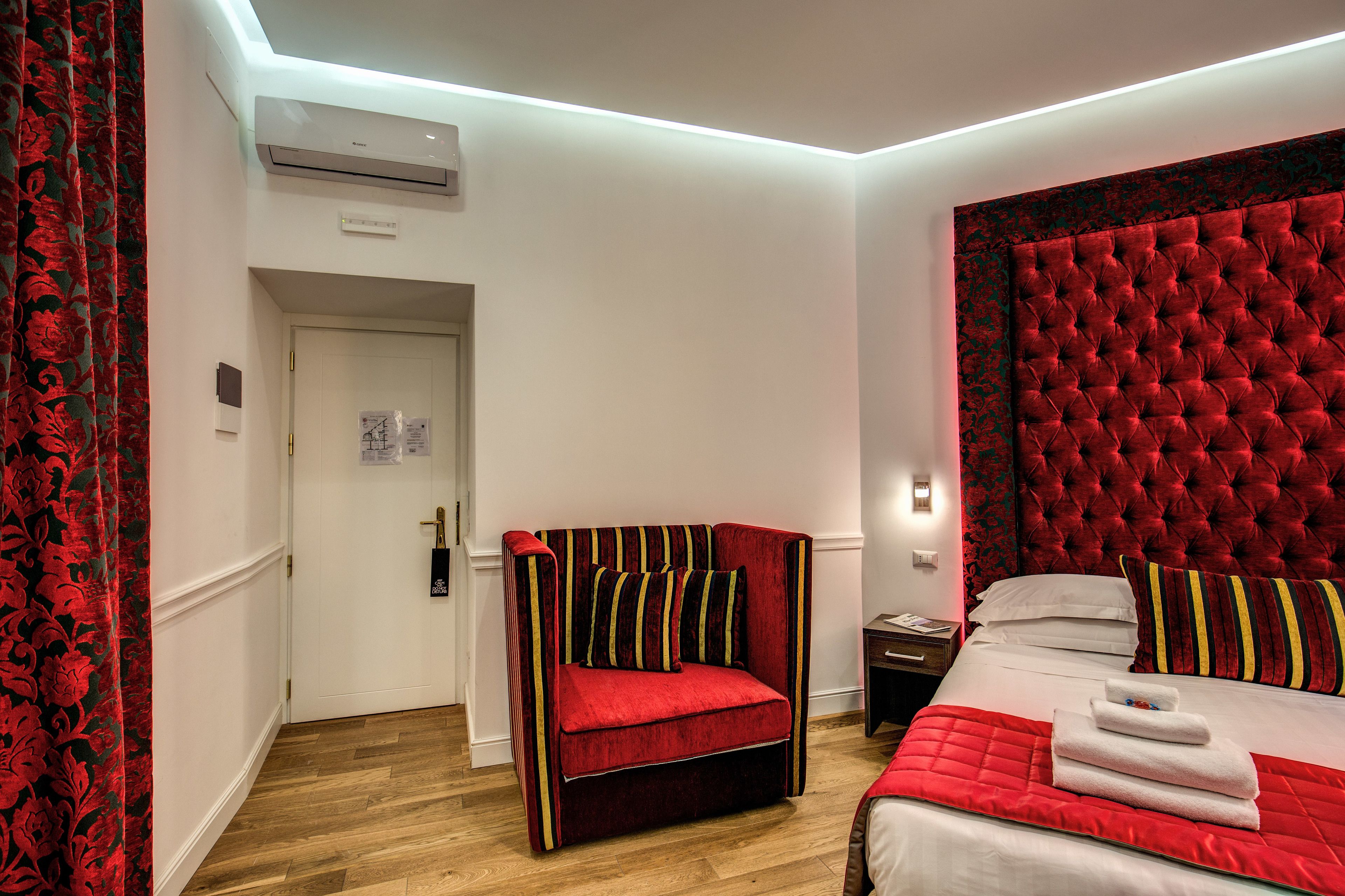 Habitación de hotel con un sillón tapizado rojo, un cabecero capitonado, una cama bien hecha con sábanas blancas y suelo de madera.