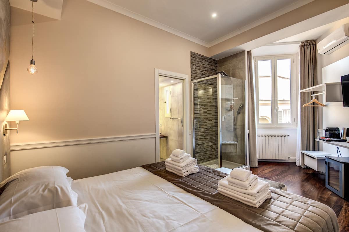 A hotel room with a bed, a bedside table, a shower area, and a desk with a computer.