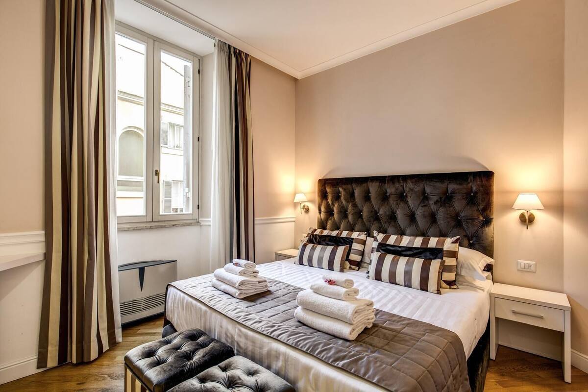 A hotel room with a large bed, a tufted headboard, two bedside tables with lamps, a window with curtains, and a bench.