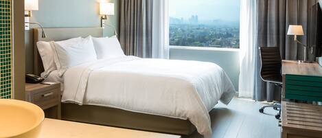 A hotel room with a bed, a desk, a chair, a TV, and a view of a cityscape.
