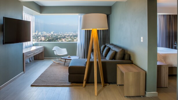 A modern hotel room with a city view, a flat-screen TV, a sofa, a lamp, and a desk. - Fiesta Inn Express Ciudad de México Forum Buenavista (Mexico City)