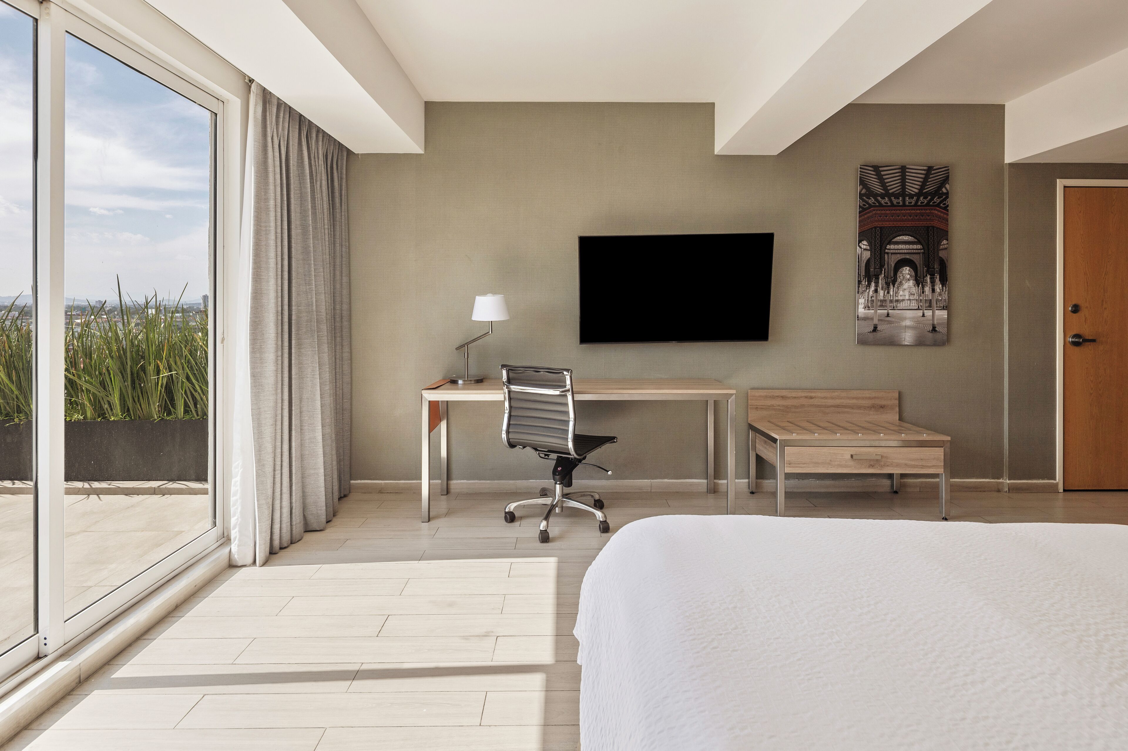 Una habitación de hotel moderna con una cama grande, un escritorio con una silla, un televisor de pantalla plana en la pared y vistas a la ciudad a través de grandes ventanales.