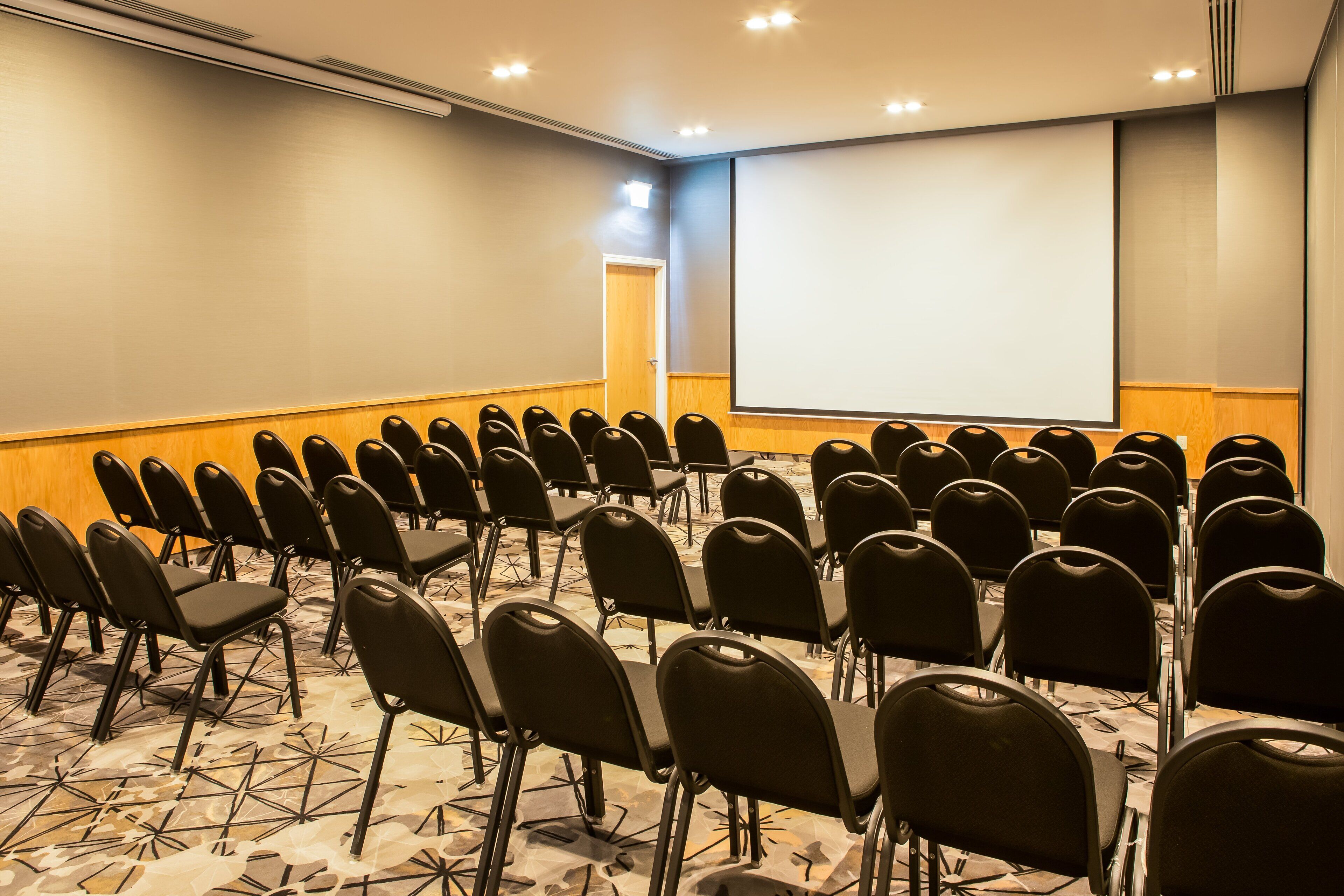 A conference room with rows of black chairs, a large screen, and a carpeted floor.