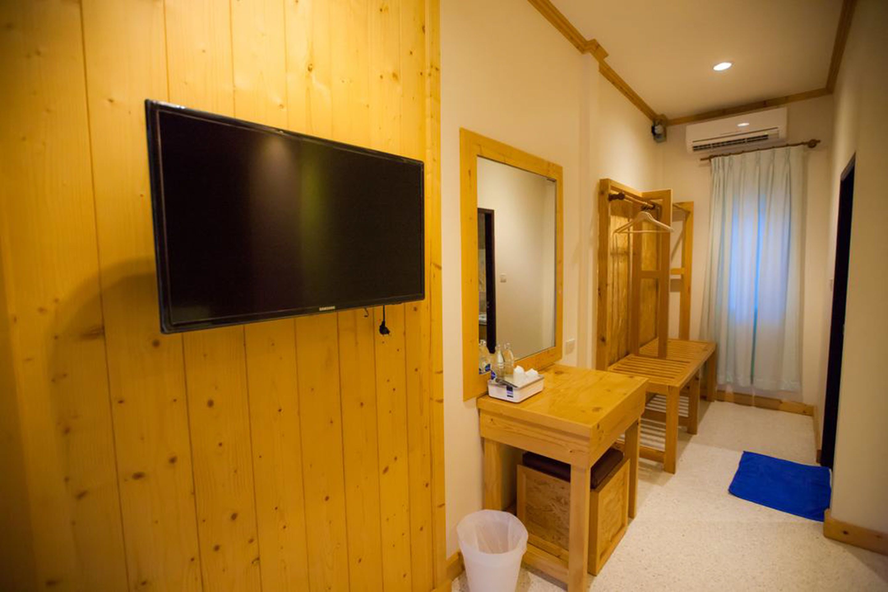 Habitación con paredes de madera, televisor montado, espejo, escritorio de madera, un banco y un cesto de basura pequeño.