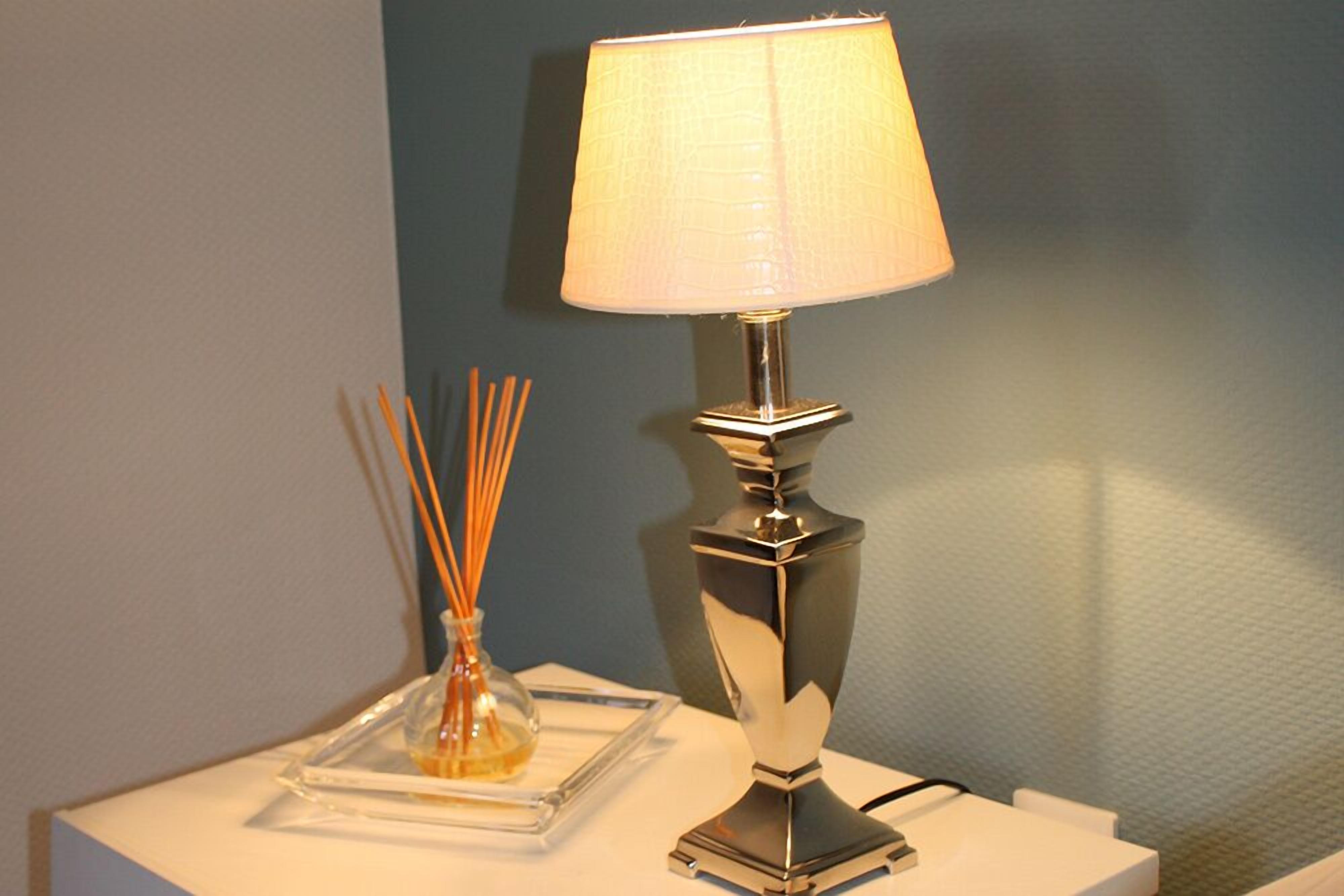 A table lamp with a square base and a cylindrical shade, placed on a white surface next to a glass vase with reed sticks.