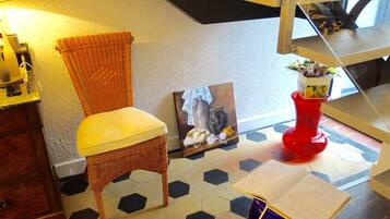 A room with a wicker chair, a painting, a red vase with flowers, and a table with a book.