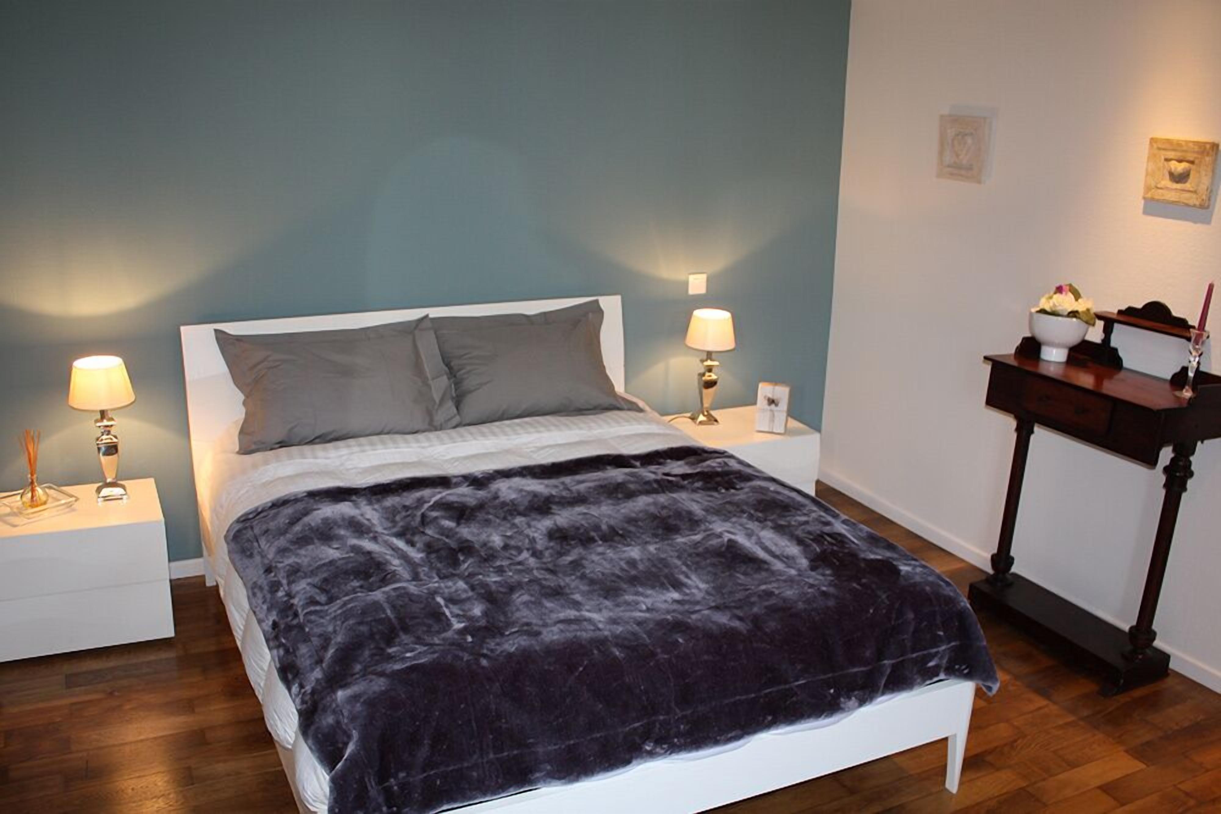 A neatly made bed with a dark blanket, two bedside lamps, and a small table with a bowl and candle.
