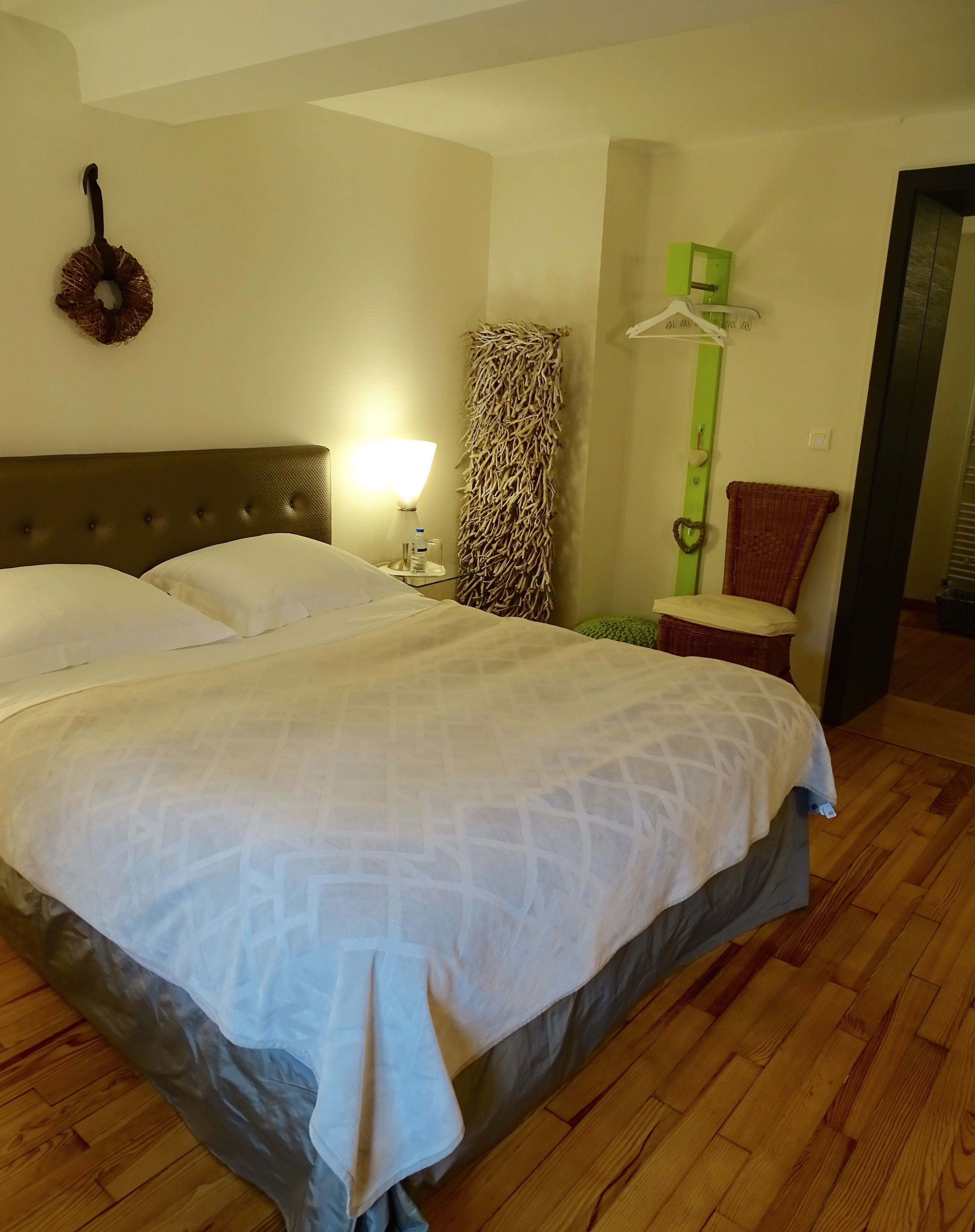 Quarto de hotel com uma cama grande, uma mesa de cabeceira com luminária, uma cadeira de vime e um ornamento verde na parede.