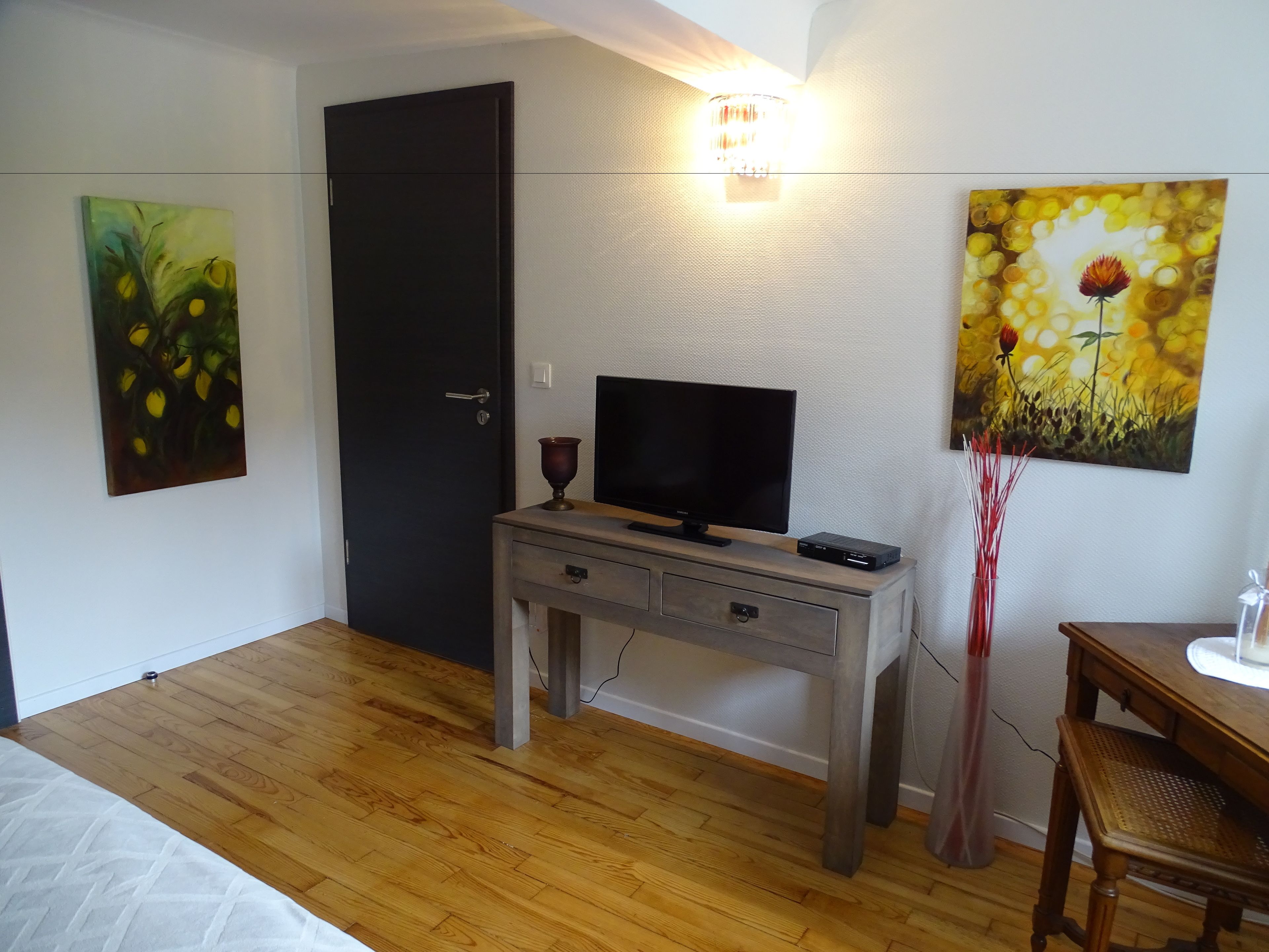 A room with a bed, a desk with a TV, a door, and two paintings on the wall.
