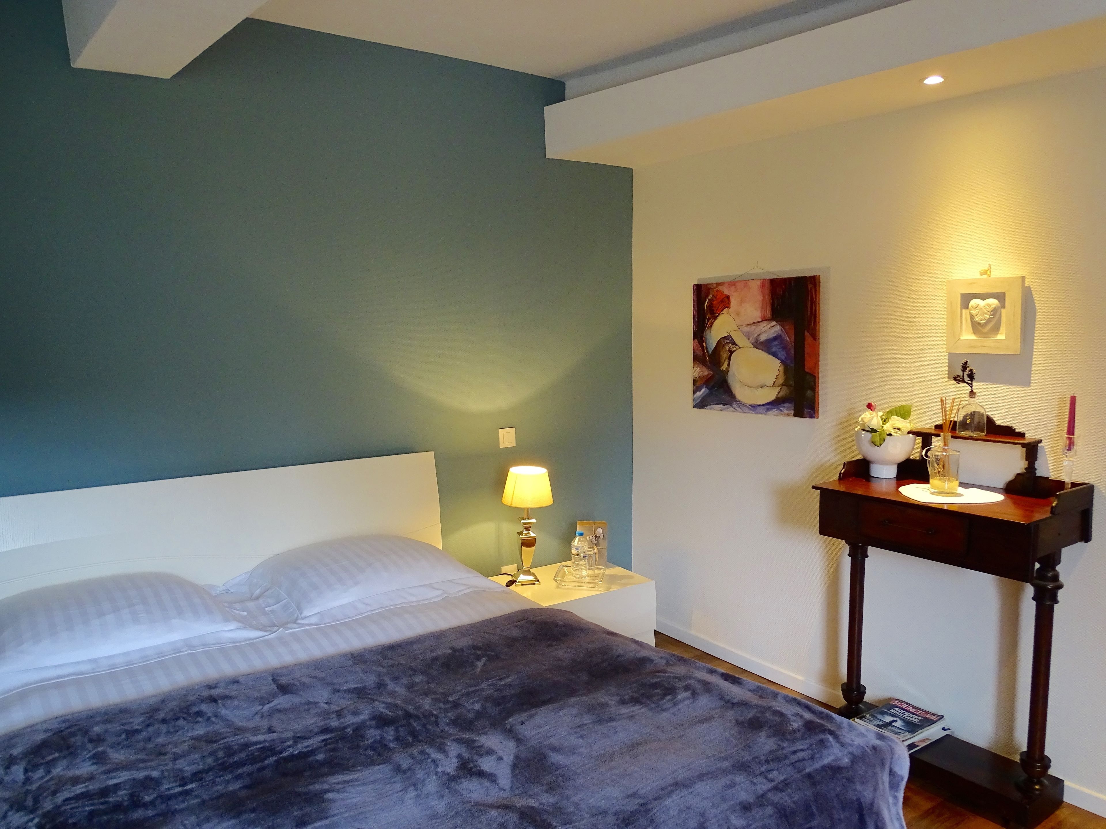 A neatly made bed with a dark blanket, a bedside lamp, a small table with a vase of flowers, and a painting on the wall.