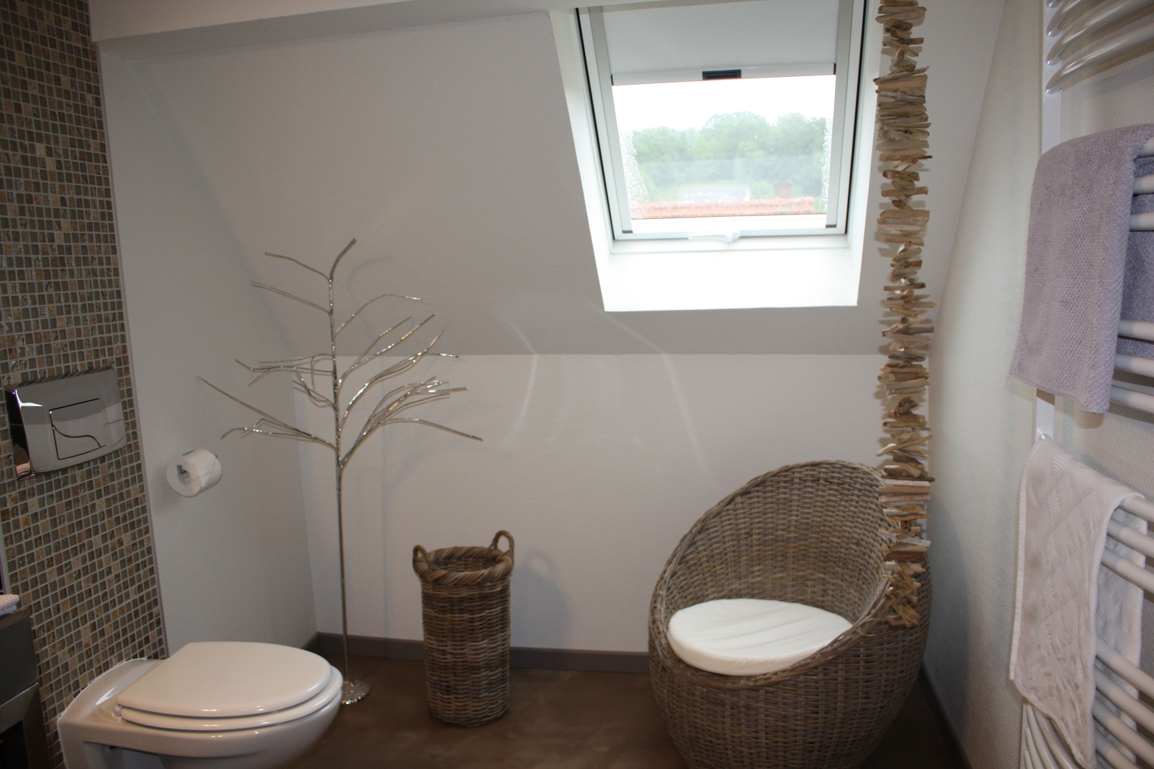 Un baño con inodoro, una silla de mimbre, una planta pequeña, una ventana y un perchero para toallas.