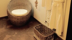 A bathroom with a wicker chair, a small wicker basket, and two white robes hanging on the wall.