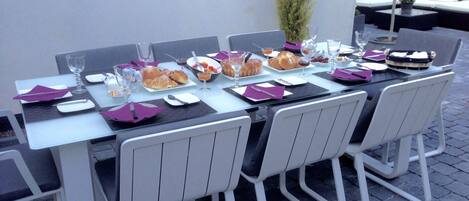 Un área de comedor al aire libre con una larga mesa dispuesta para una comida, rodeada de sillas blancas.