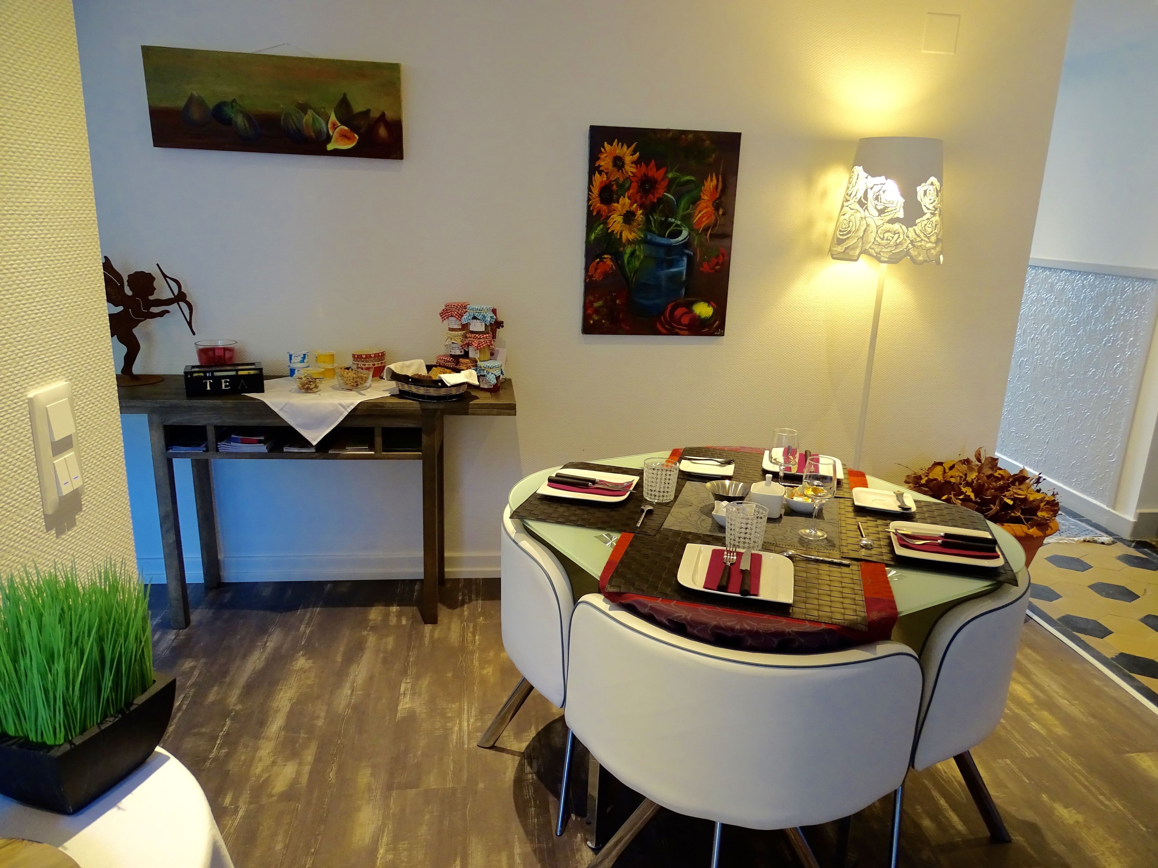 Un comedor con una mesa dispuesta para cuatro, una pequeña cocina con estación de té y una pared decorada con dos cuadros.