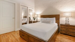 Een slaapkamer met een houten bed, witte muren, een accentmuur van baksteen, een witte deur en een nachtkastje met een lamp.